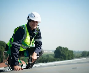 A worker wearing a white helmet, safety harness, and reflective vest kneels on a rooftop, delivering expert commercial roofing services in The Woodlands. The background shows trees and a clear sky.