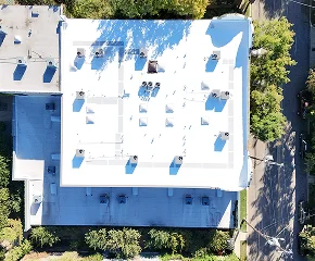 Aerial view of a white flat-roofed building with multiple vents and equipment on top, surrounded by trees and adjacent streets in The Woodlands—an example where Noble Roofing provides trusted roofing services.