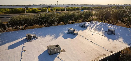 A flat white commercial building roof with several HVAC units, expertly handled by Noble Roofing, is surrounded by trees, with a highway and overpass in the background under a clear sky.