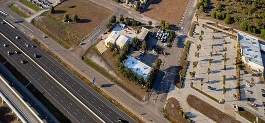Aerial view of a highway with several lanes of traffic next to buildings, parking lots, and green spaces with scattered trees in The Woodlands, where Noble Roofing offers trusted roofing services.