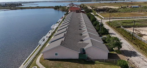 Aerial view of a row of waterfront houses with gray Noble Roofing-installed roofs beside a body of water, bordered by a road and grassy fields on the other side.