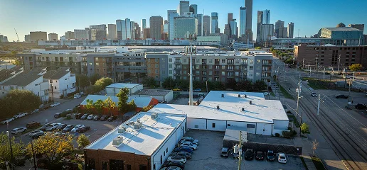 Aerial view of The Woodlands cityscape with tall buildings in the background, mid-rise apartments, parked cars, and white-roofed warehouses—showcasing quality commercial roofing—under a clear blue sky.