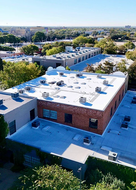 Aerial view of flat-roofed commercial buildings in The Woodlands with white rooftops equipped with multiple HVAC units, showcasing quality commercial roofing by Noble Roofing, surrounded by trees and greenery under a clear sky.