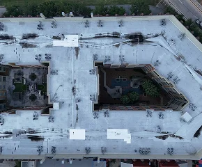 Aerial view of a large, light-colored rooftop with an irregular U-shaped design by Noble Roofing, surrounding a central courtyard with greenery and small structures. Dark streaks and vents are visible on the roof’s surface, showcasing expert commercial roofing.
