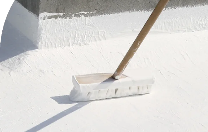 A brush with a wooden handle is being used to apply a thick layer of white coating to a flat surface near a wall, showcasing commercial roofing work by Noble Roofing in The Woodlands.