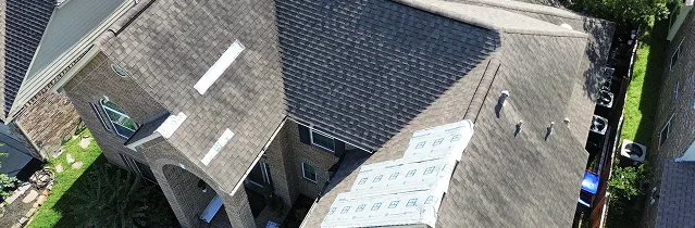 Aerial view of a large brick house in The Woodlands with a multi-sectioned roof; part features new shingles installed by Noble Roofing, while another section is under repair. Neighboring houses and a yard are partially visible.