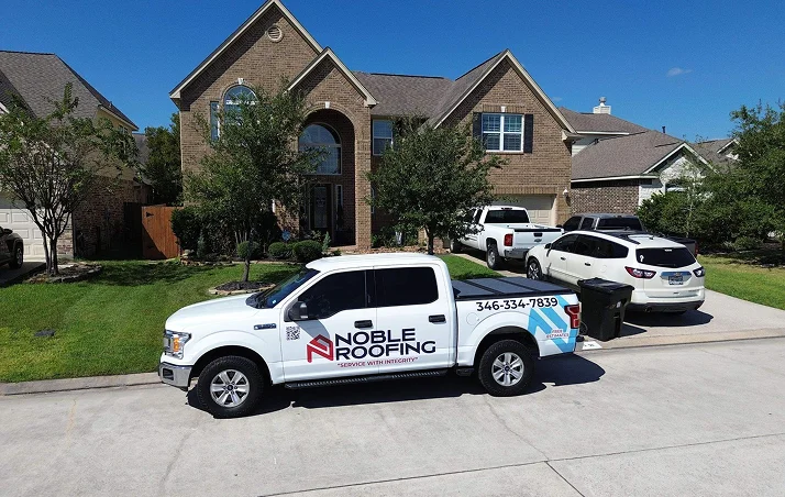A white pickup truck with Noble Roofing branding, showcasing their expert roofing services, is parked on the street in front of a large brick house with a well-kept lawn and several other vehicles in the driveway on a sunny day.