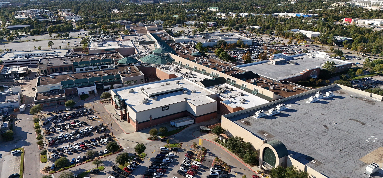 Aerial view of The Woodlands shopping mall complex, surrounded by parking lots filled with cars, green trees, and additional buildings—showcasing prominent commercial roofing by Noble Roofing.