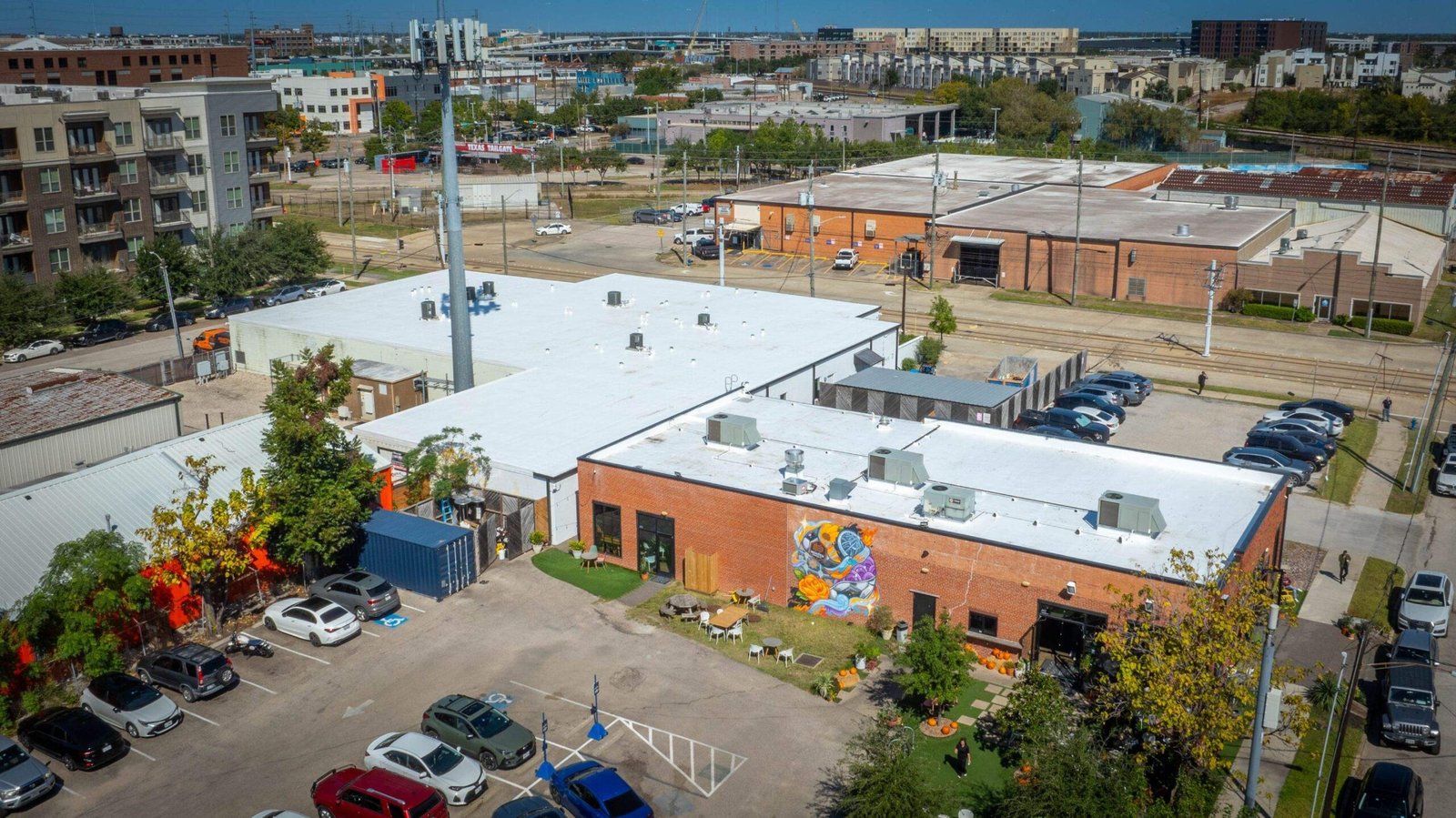 Aerial view of a parking lot surrounded by brick and metal buildings, some serviced by Noble Roofing, with parked cars, a mural on one wall, and city structures in the background under a clear sky.