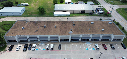 Aerial view of a single-story commercial building with multiple storefronts, parking spaces, and several cars parked in front. Noble Roofing offers expert commercial roofing services for businesses in this area surrounded by grassy lots, roads, and warehouses.