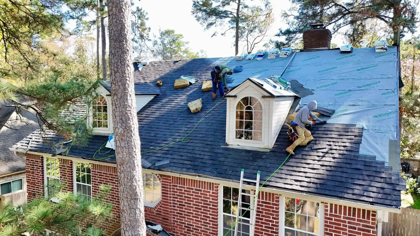 Several workers from Noble Roofing are installing new shingles and underlayment on the roof of a brick house in The Woodlands. Roofing materials and tools are scattered atop the structure, with a ladder leaning against the house.