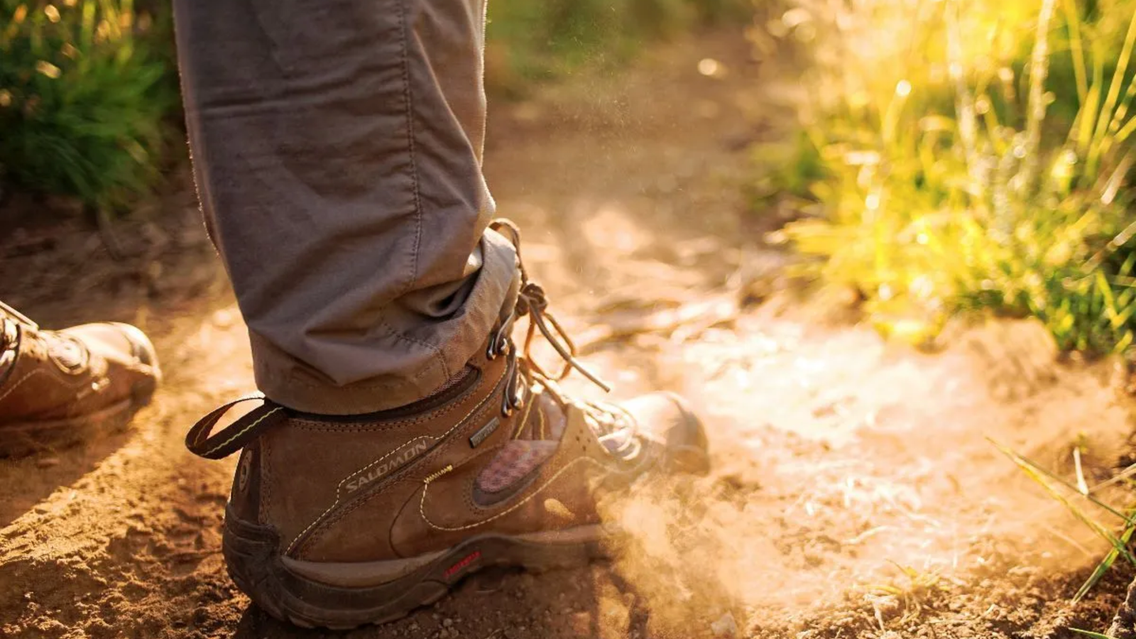 A close-up of a person’s feet in brown hiking boots and khaki pants, stepping on a dusty sunlit trail in The Woodlands, with dry soil and grass visible in warm, golden light—capturing the spirit of adventure Noble Roofing brings to commercial roofing projects.