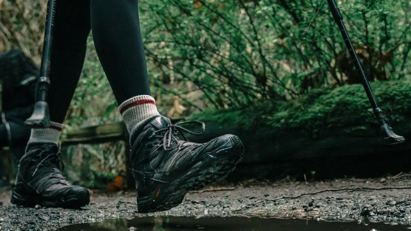 Close-up of a person’s legs wearing black hiking boots and gray socks, walking with trekking poles through a muddy, forested trail in The Woodlands with lush green foliage in the background.