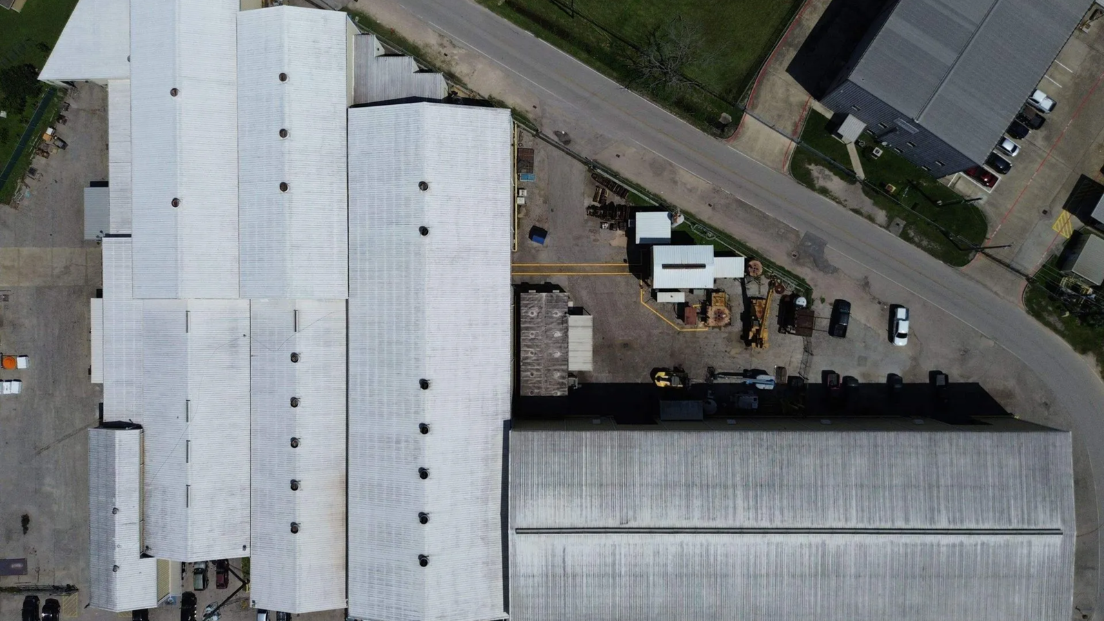 Aerial view of large industrial buildings with white roofs showcases Noble Roofing’s expert commercial roofing services. Smaller structures, parked cars, and equipment surround the site, bordered by grass and pavement.