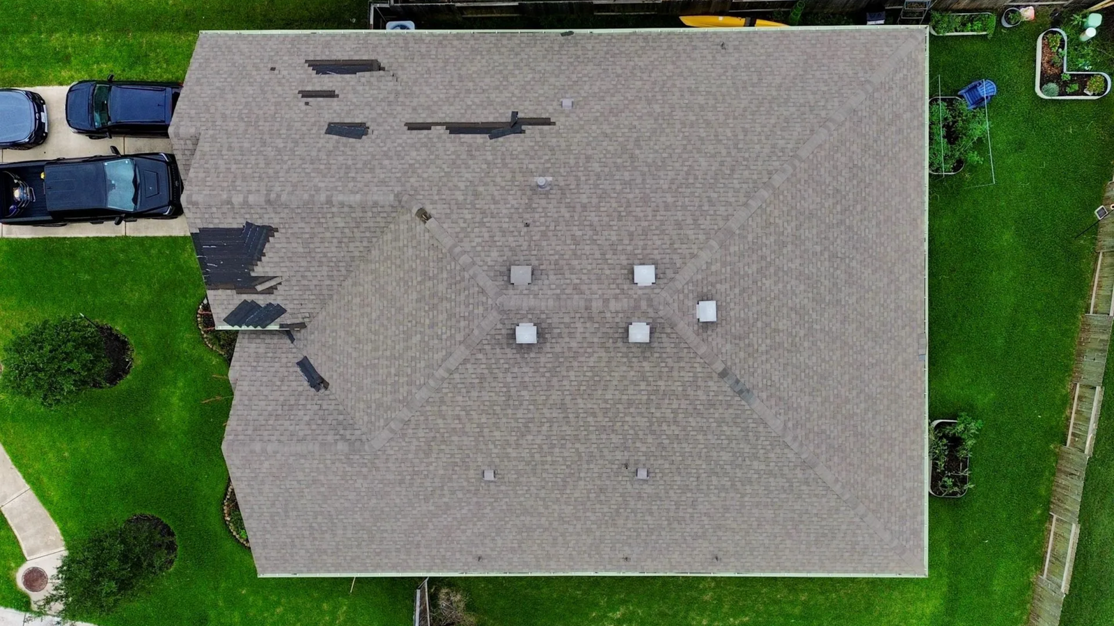 Aerial view of a house roof in The Woodlands with missing and damaged shingles in several areas. Surrounding the house is green grass, a driveway with parked cars, and parts of a fenced yard—ideal for Noble Roofing’s expert assessment.