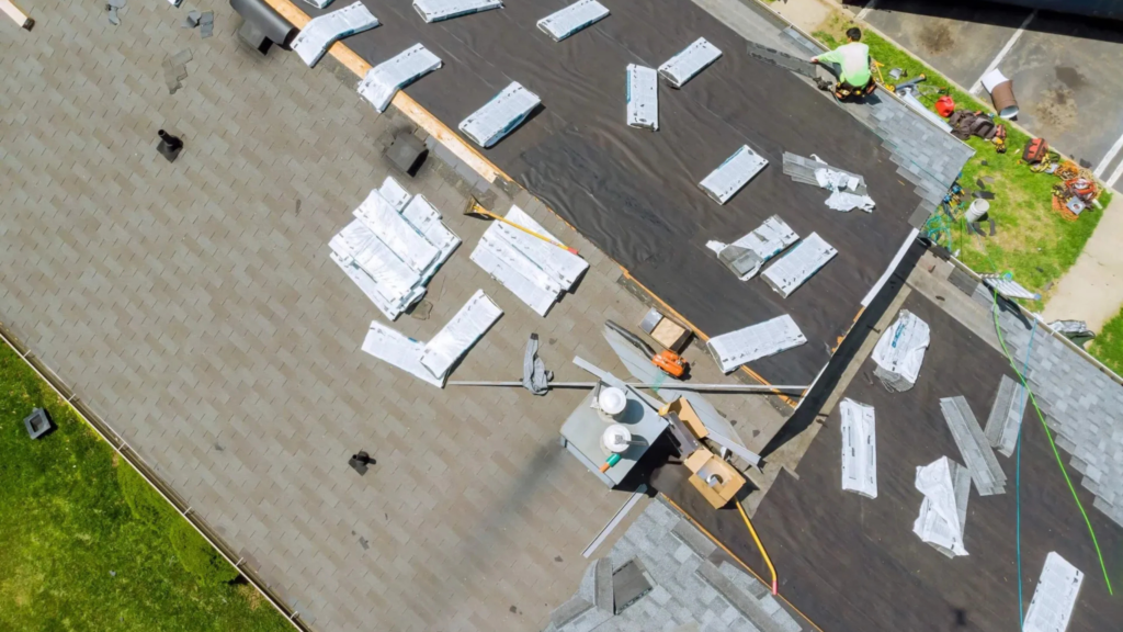 Aerial view of a house roof under construction in The Woodlands, with roofing materials and tools scattered on top. Noble Roofing workers, equipped with safety gear, are installing tar paper as part of their expert roofing services.