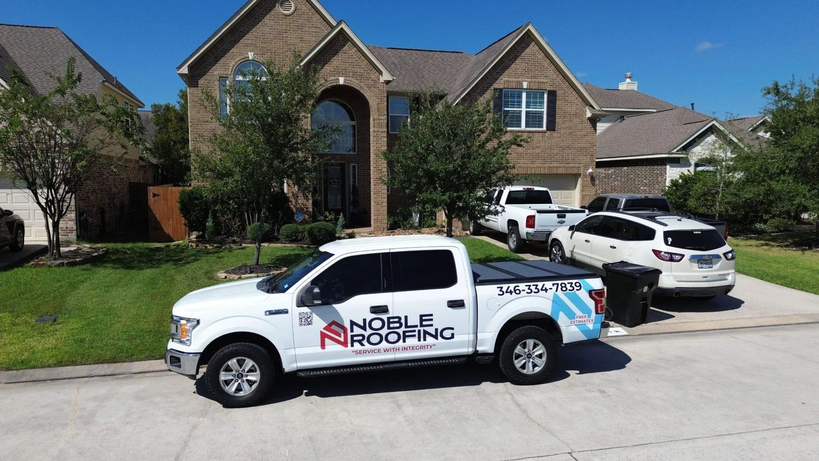 A white Noble Roofing truck is parked in front of a two-story brick house with a well-kept lawn. Several cars are in the driveway and on the street, highlighting Noble Roofing’s trusted roofing services. Trees and blue sky complete the scene.