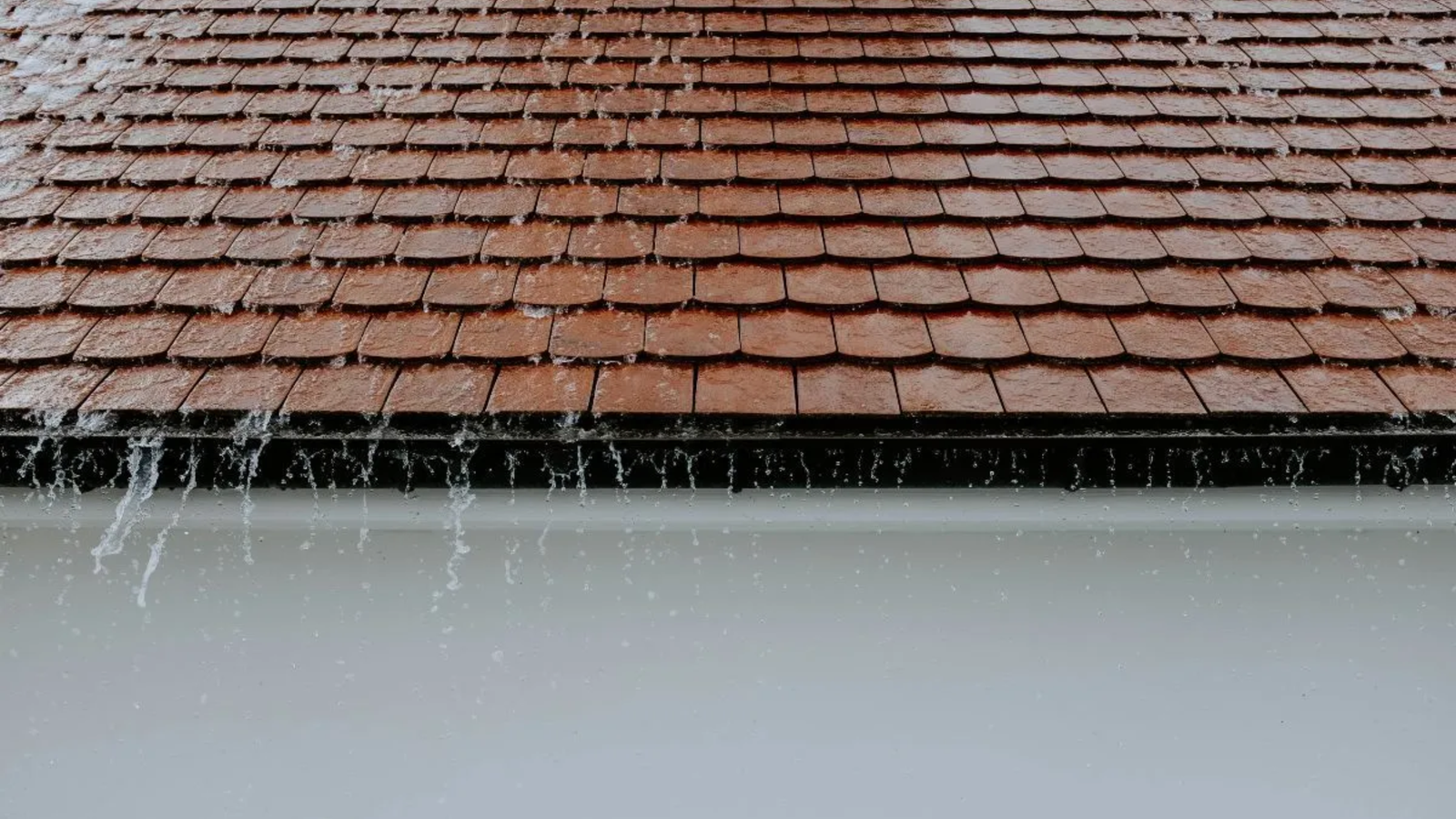 Red roof tiles with rainwater streaming off the edges, creating visible flows of water against a white wall. The image captures a rainy day in The Woodlands and highlights the importance of quality roofing services for proper drainage.