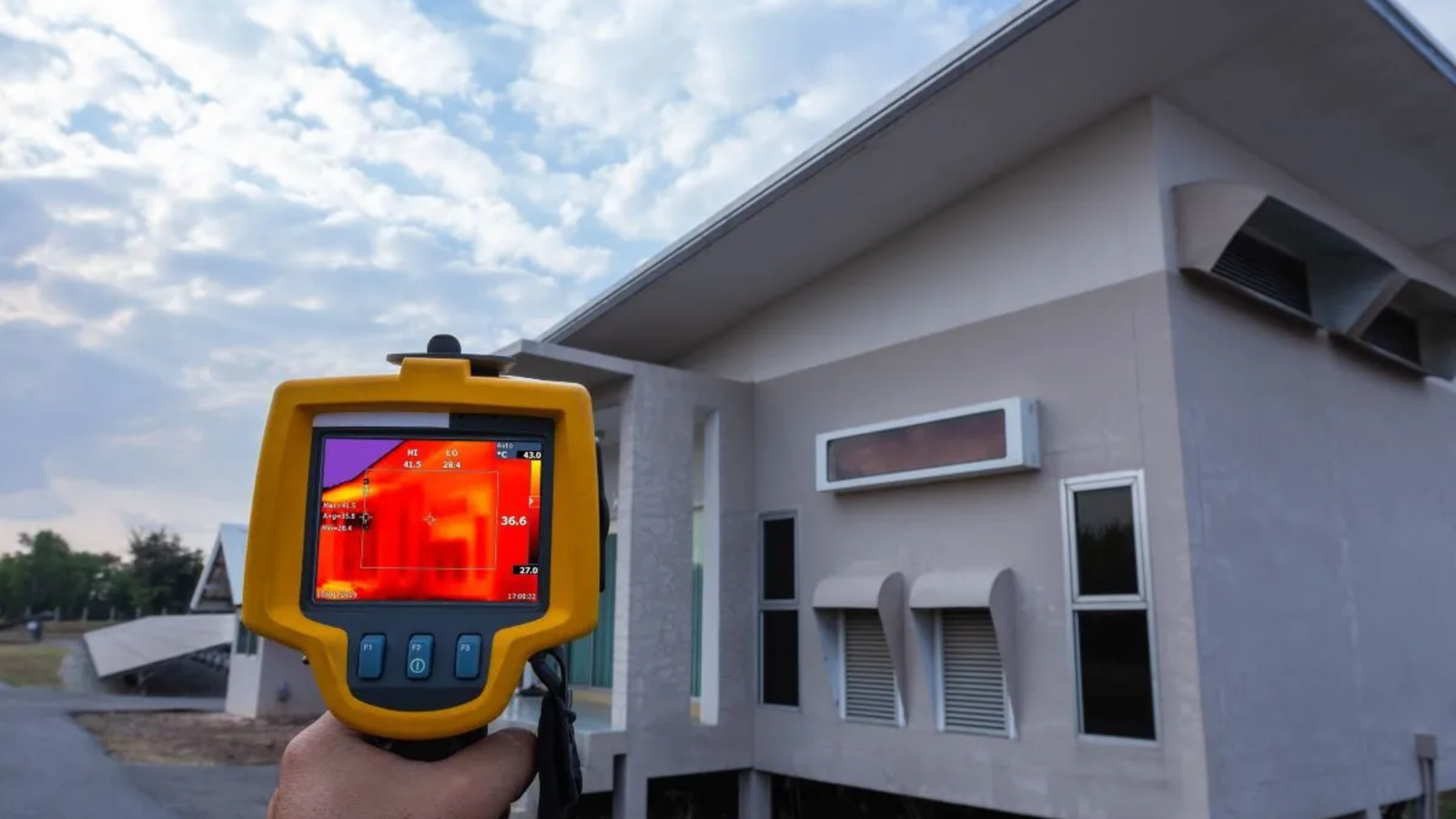 A person holds a thermal imaging camera pointed at a modern house with a sloped roof, highlighting heat levels in red and yellow. The pale gray exterior features unique windows—perfect for showcasing Noble Roofing’s expert roofing services.