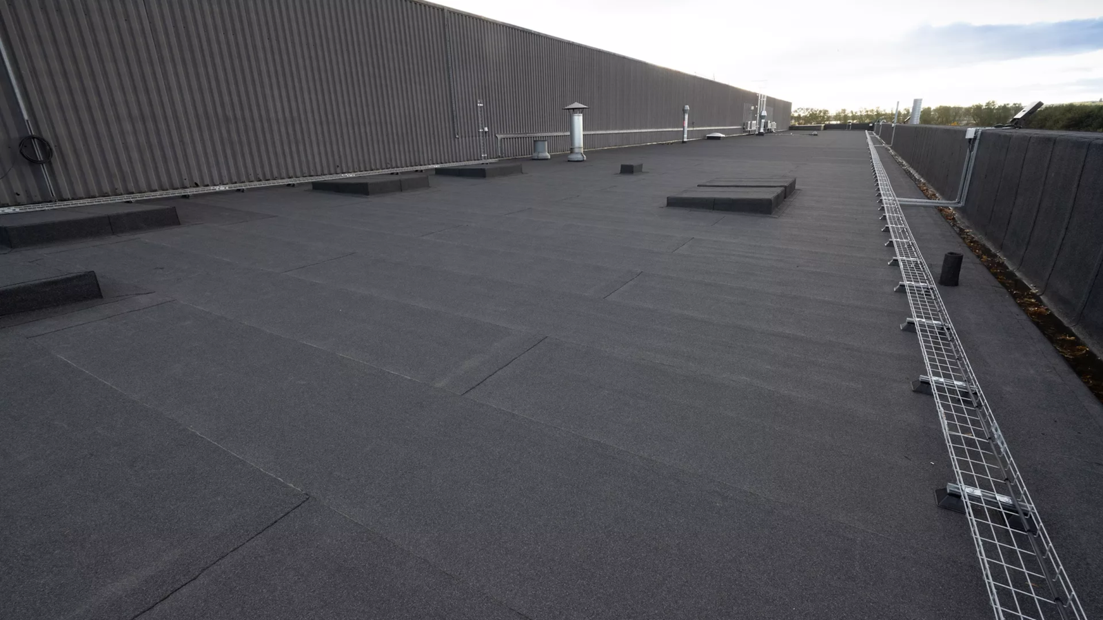 Flat commercial building roof covered with dark roofing material by Noble Roofing, featuring rectangular vents and raised areas; metal safety railing and piping run along the edge. Reliable roofing services available in The Woodlands.