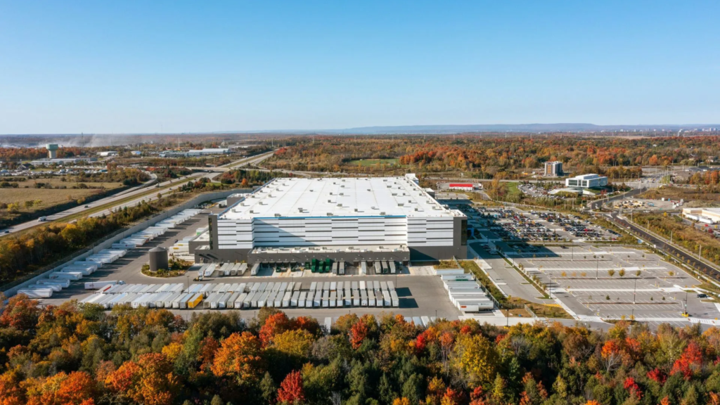 A large industrial warehouse or distribution center in The Woodlands, serviced by Noble Roofing, is surrounded by parking lots, loading docks, and autumn-colored trees under a clear blue sky. Roads and distant buildings are visible in the background.