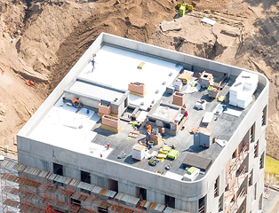 Aerial view of a building under construction in The Woodlands, showing workers and equipment from Noble Roofing on the flat rooftop, surrounded by scaffolding and dirt at the commercial roofing site.