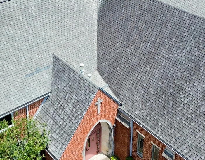 Aerial view of a brick church building in The Woodlands with gray shingle roofing, a white cross above the entrance, and pink double doors framed by stonework. Some greenery is visible near the entrance, showcasing quality commercial roofing.