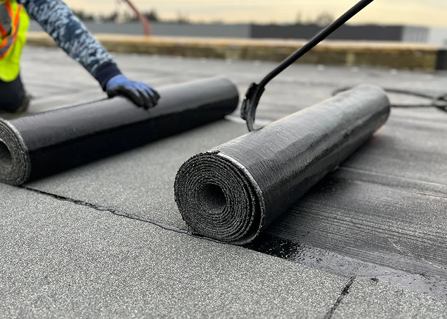 A worker in safety gear is installing roll roofing material on a flat roof for Noble Roofing in The Woodlands, unrolling a sheet while another roll lies beside it. The surface appears freshly coated with adhesive, showcasing expert roofing services.