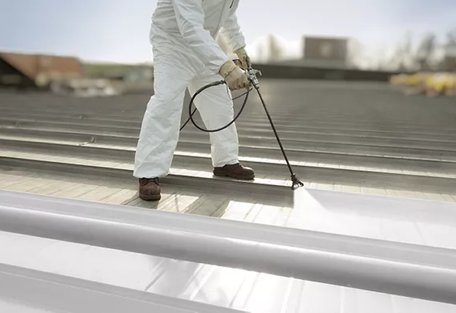 A person wearing a white protective suit and brown shoes uses a spray gun to apply coating or paint to the surface of a metal roof outdoors—showcasing professional roofing services by Noble Roofing in The Woodlands.