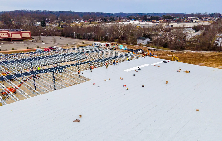 Aerial view of Noble Roofing construction workers assembling the roof of a large commercial building in The Woodlands, with metal framework exposed on one side and completed commercial roofing on the other, surrounded by equipment and a parking lot.