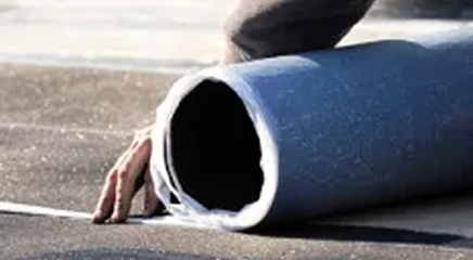 A person’s hand is unrolling a sheet of roofing material onto a flat surface, possibly preparing or repairing a roof as part of professional Roofing Services.