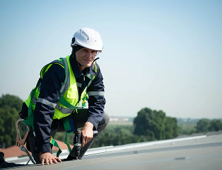 A construction worker wearing a white helmet, safety vest, and harness kneels on a rooftop, smiling at the camera—representing Noble Roofing’s commitment to quality commercial roofing, with trees and buildings visible in the blurred background.