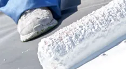 A close-up of a white paint roller applying white coating on a surface, with a person’s foot in a white sneaker and blue pants visible in the background—perfect for showcasing Noble Roofing’s expert commercial roofing services.