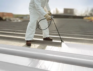 A person in protective clothing sprays a coating onto a metal roof using a spray gun, likely applying paint or sealant for commercial roofing in The Woodlands with Noble Roofing.