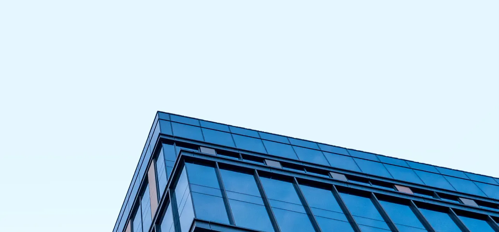 A close-up, angled view of the corner of a modern glass office building with blue-tinted windows against a clear, light blue sky showcases the premium work by Noble Roofing, experts in commercial roofing solutions.