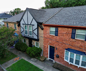 A two-story townhouse in The Woodlands features a mix of brick and Tudor-style siding, blue doors and windows. The sidewalk, bushes, and a patch of grass are visible in front—expertly maintained by Noble Roofing’s trusted roofing services.