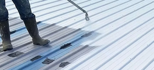 A person wearing boots is spraying a white coating onto a corrugated metal roof, covering dark patches and creating a cleaner, uniform surface—showcasing the quality of Noble Roofing’s commercial roofing services.