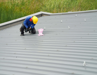 A worker in a yellow helmet and blue shirt kneels on a metal roof, using tools near a pink object, with green vegetation behind—illustrating the dedication of Noble Roofing providing expert roofing services in The Woodlands.
