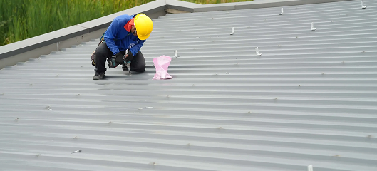 A worker in a yellow hard hat and blue shirt kneels on a metal roof, using a tool near a pink plastic bag, with green vegetation in the background—a typical scene during Noble Roofing’s commercial roofing services.