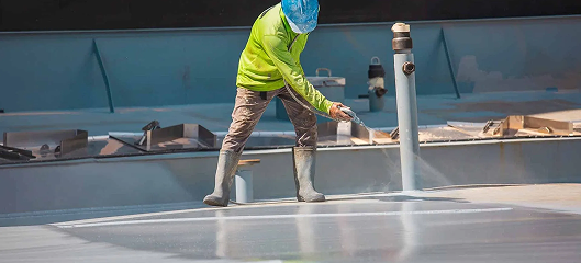 A worker wearing a blue hard hat, green shirt, and boots sprays waterproof coating onto a large flat surface, showcasing Noble Roofing’s expert commercial roofing services for construction or maintenance projects.