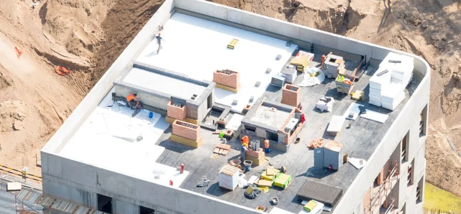 Aerial view of several workers and construction materials on the roof of a rectangular building under construction in The Woodlands, with Noble Roofing providing expert roofing services amid dirt and sand piles.