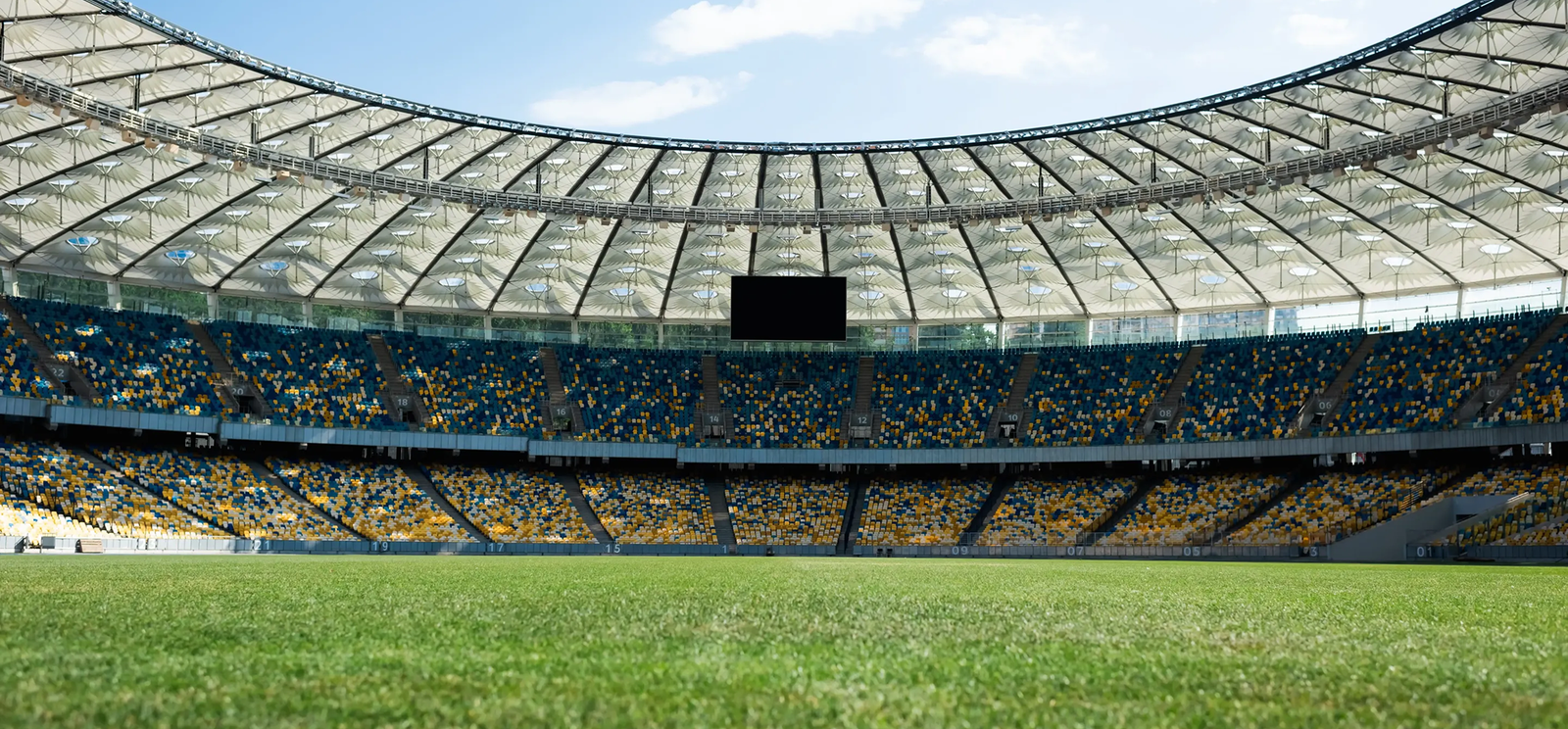 A large, empty sports stadium in The Woodlands features rows of blue and yellow seats, a green grass field, and a partially transparent roof—showcasing the quality craftsmanship found in Noble Roofing’s roofing services.
