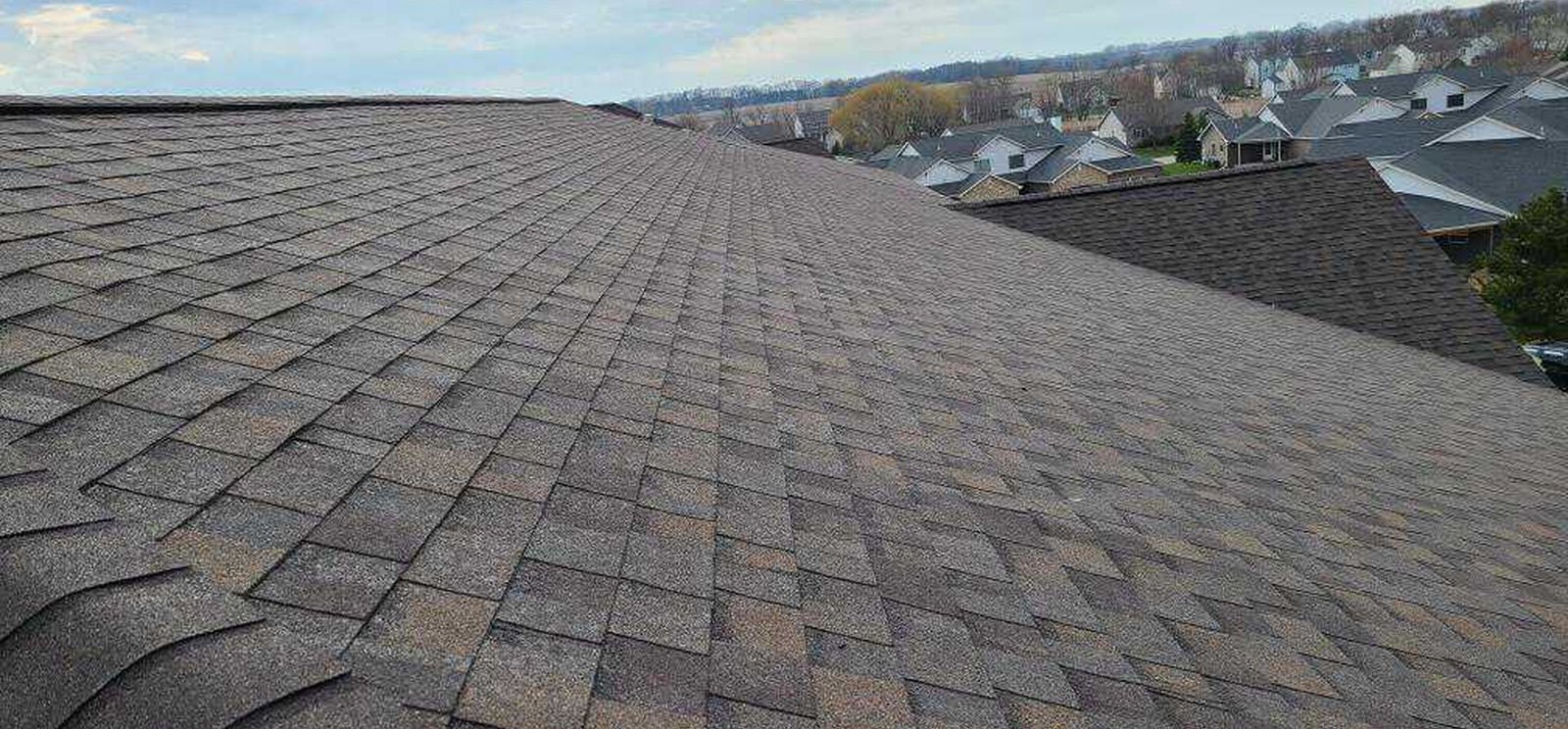 View from a rooftop covered with brown asphalt shingles, overlooking The Woodlands neighborhood with houses and trees in the distance under a partly cloudy sky—an ideal spot to appreciate quality roofing services.