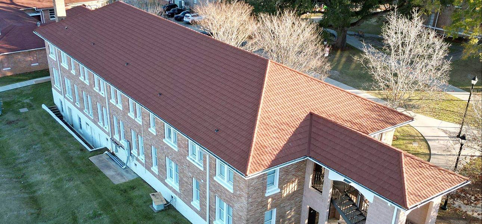 Aerial view of a large, two-story brick building with a red gabled roof by Noble Roofing, surrounded by grass, trees, sidewalks, and adjacent parking area in The Woodlands—showcasing expert commercial roofing.