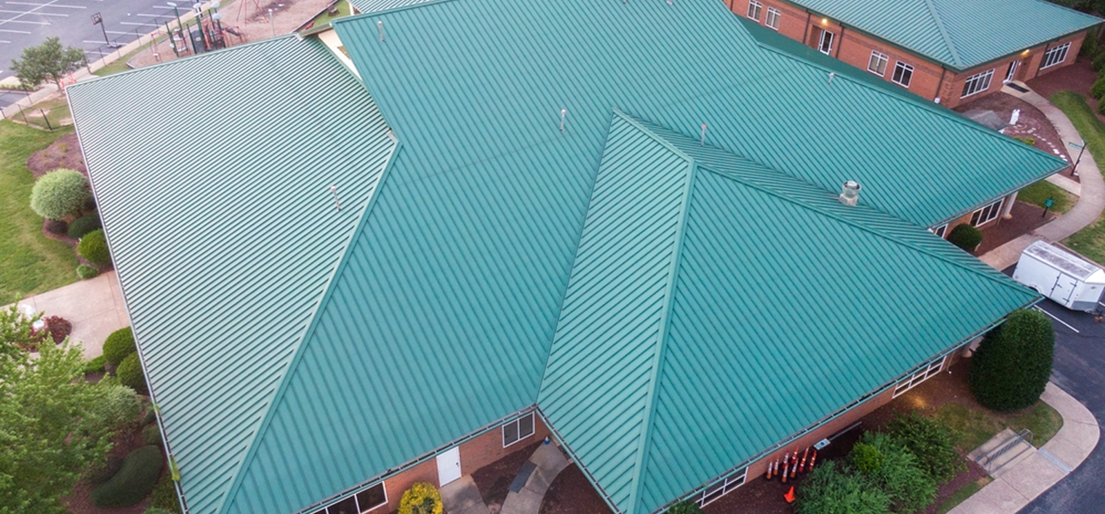Aerial view of a large building with a green metal roof featuring multiple peaks and angles, showcasing expert commercial roofing in The Woodlands, surrounded by landscaped greenery and walkways.