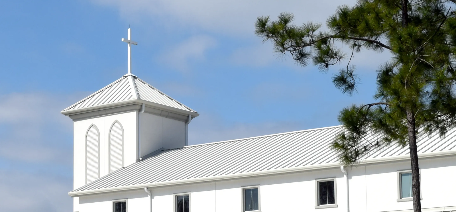 A white church with a metal roof installed by Noble Roofing and a steeple topped by a cross, set against a blue sky with clouds and a pine tree on the right side.