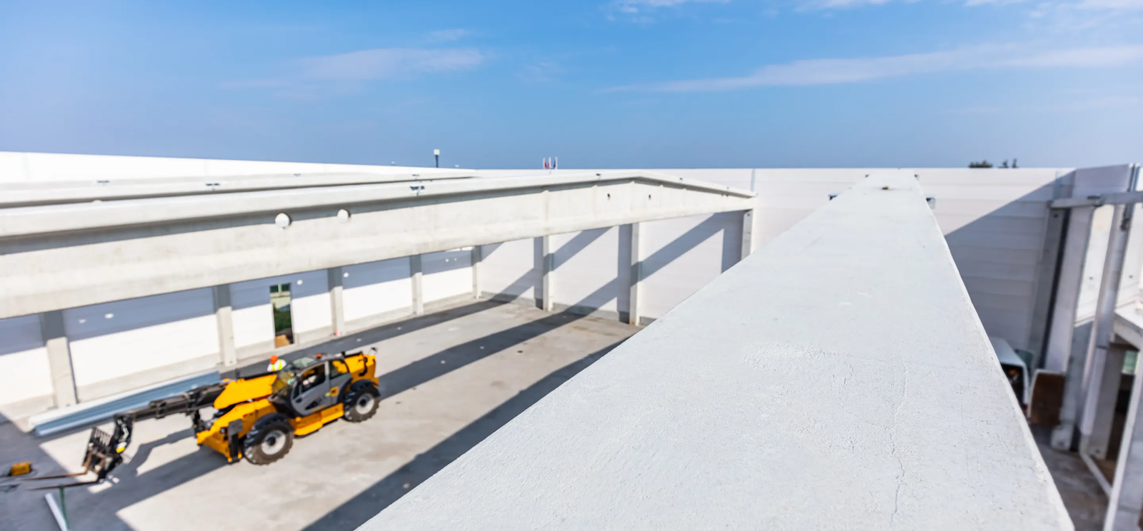 A modern industrial building under construction, featuring concrete walls and beams, showcases Noble Roofing's expertise in commercial roofing, with a yellow telescopic forklift parked in the open courtyard under a clear blue sky.