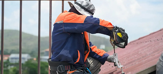 A worker in a safety harness and hat uses a power tool on a sloped metal roof, showcasing professional commercial roofing services in The Woodlands, with buildings and green hills visible in the background.