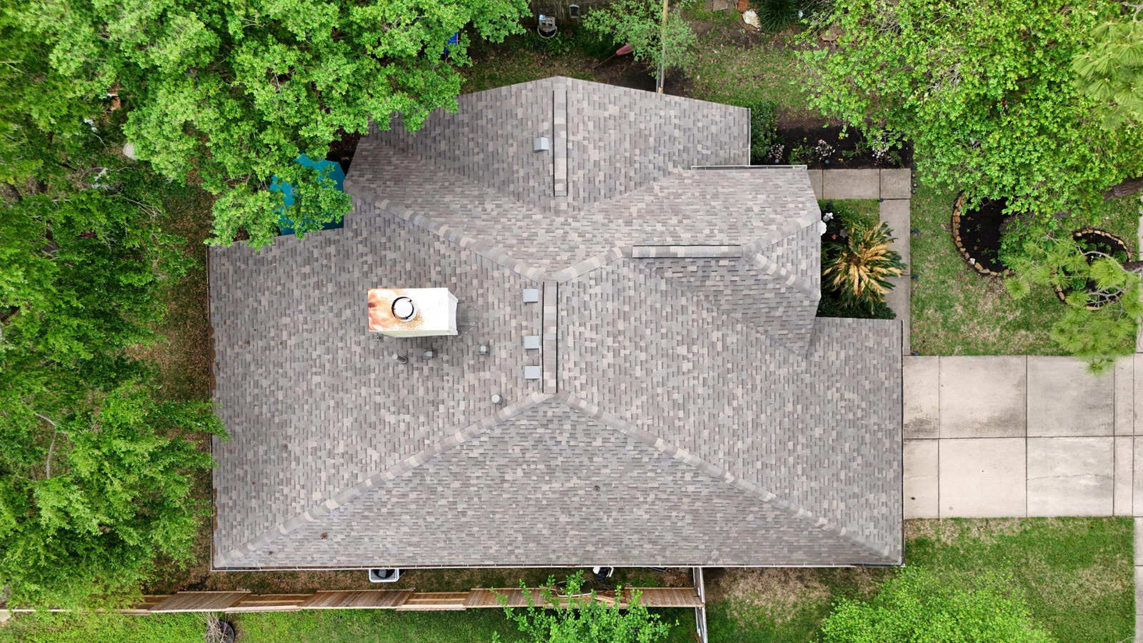 Aerial view of a gray-shingled house roof in The Woodlands, surrounded by green trees, with a concrete driveway on the right and a wooden fence below. The roof has a chimney and multiple ridges—ideal for showcasing Roofing Services.