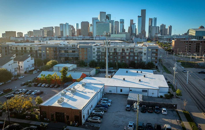 Aerial view of a cityscape with modern high-rise buildings, mid-rise apartments, parking lots, and low-rise buildings in the foreground under a clear blue sky—an impressive backdrop for Noble Roofing’s expert commercial roofing services.